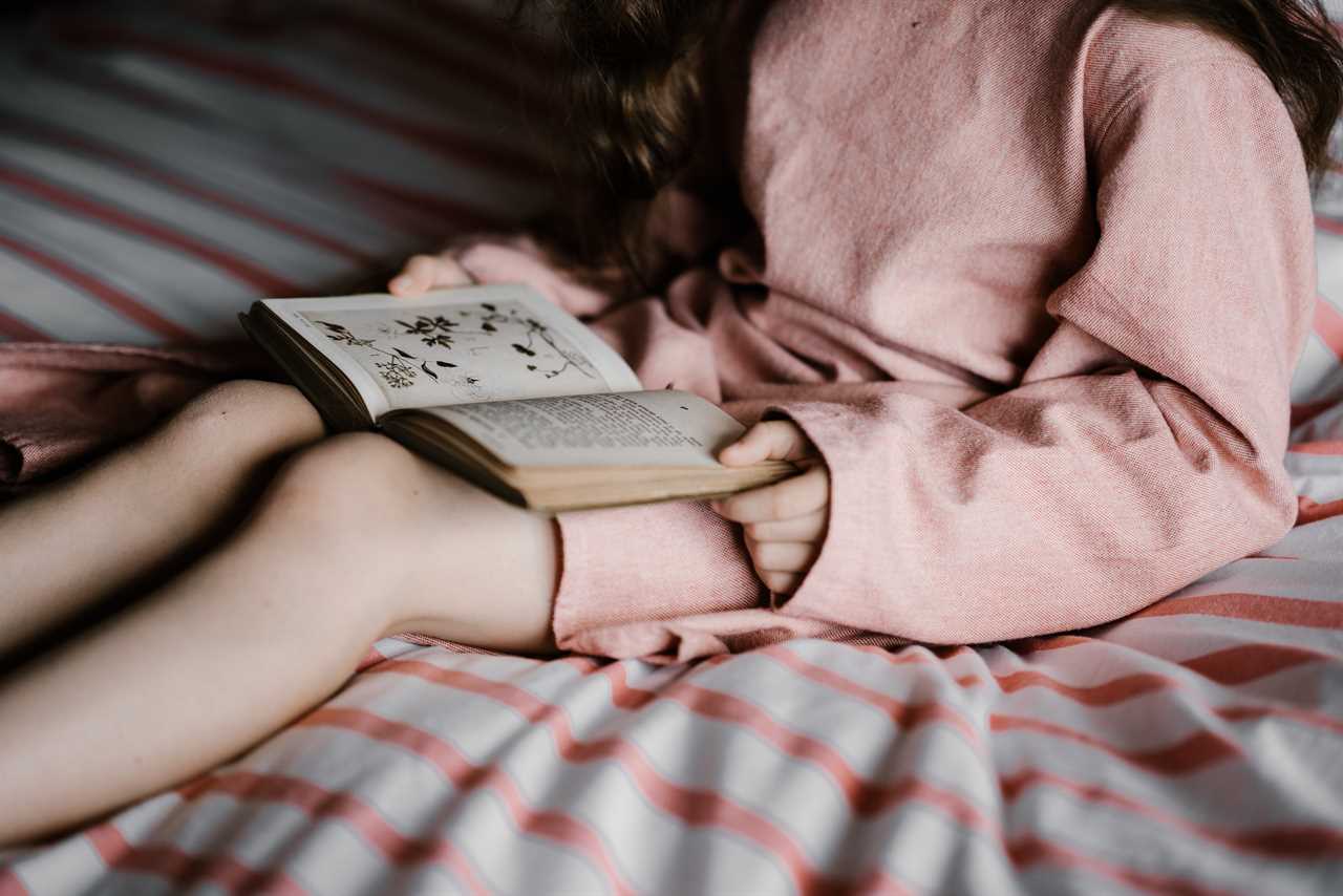 A girl sitting on a bed reading a book.