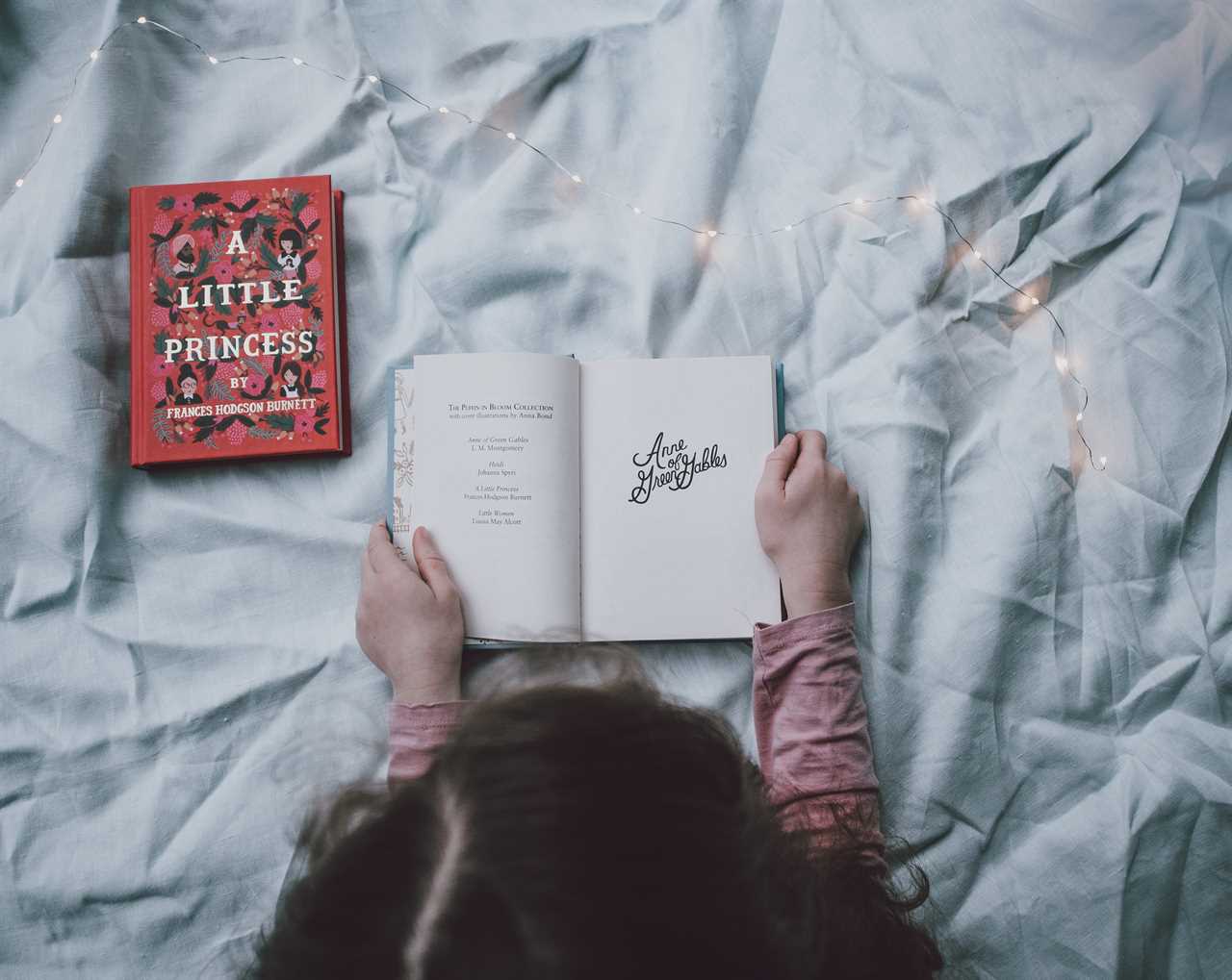 A girl is reading a book on a bed with lights around it.