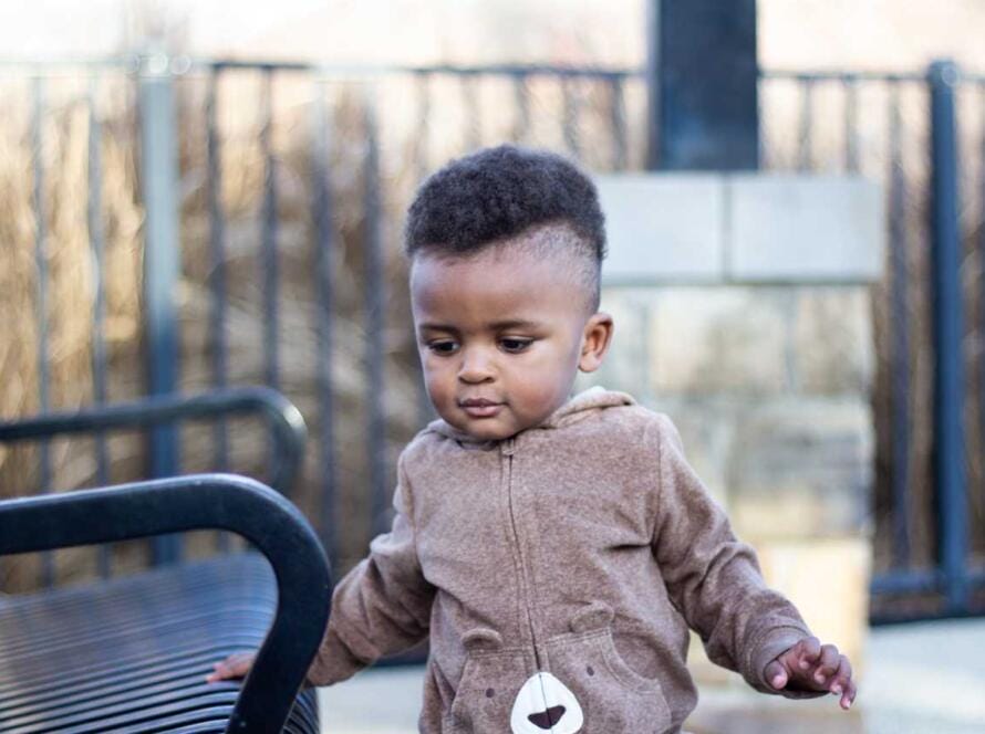 A baby boy in a brown bear outfit walking on a bench.