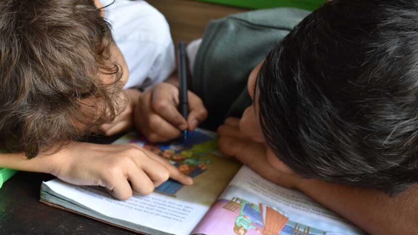 Two boys are sitting at a table reading a book.