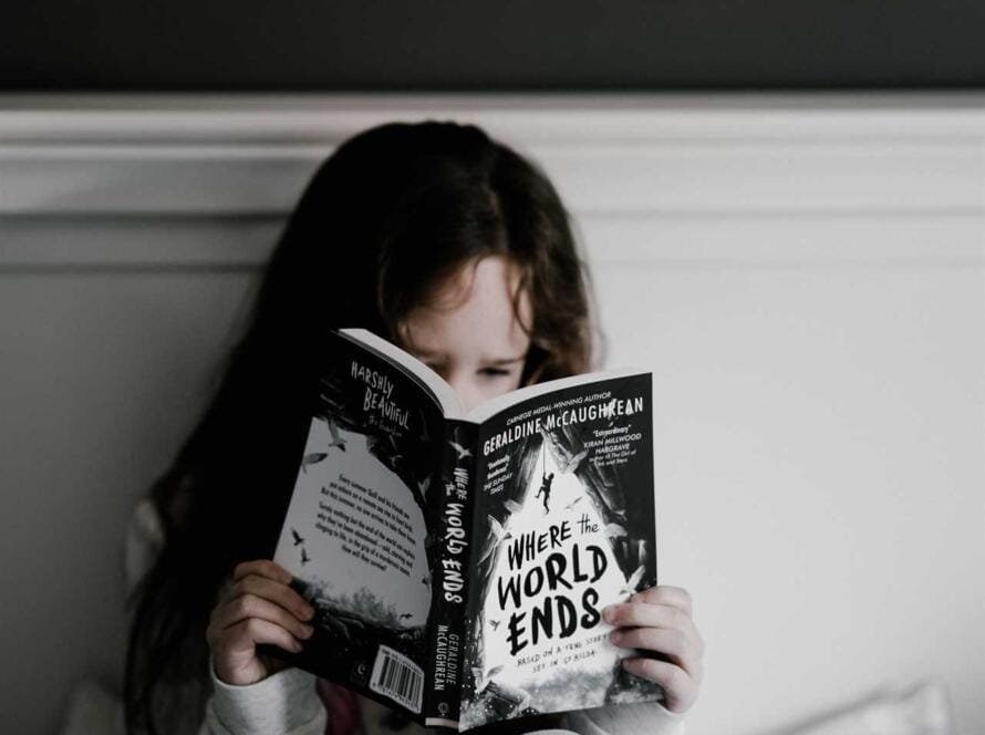 A girl sitting on a bed reading a book.