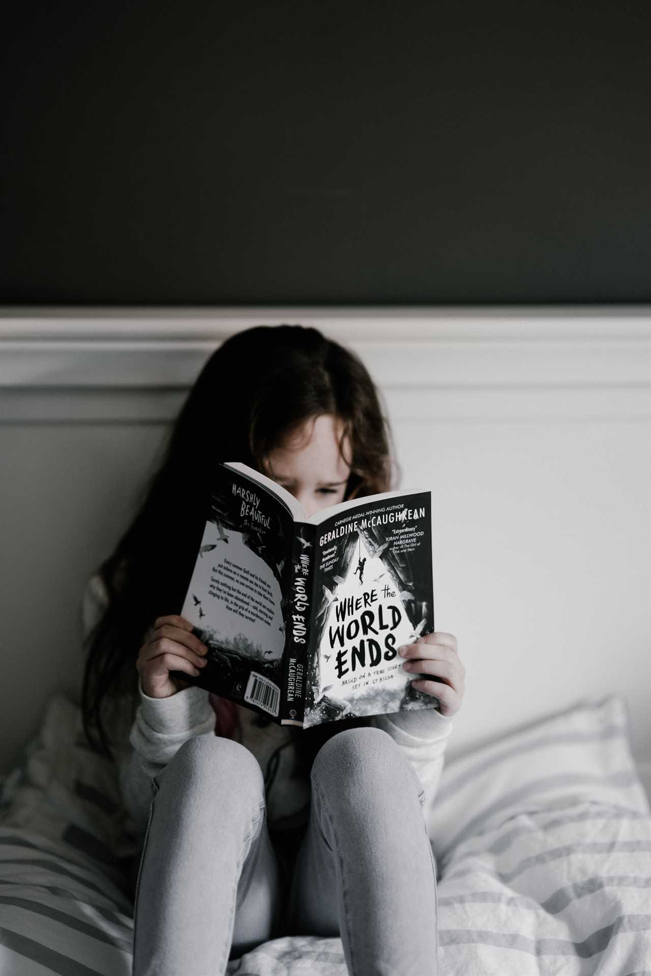 A girl sitting on a bed reading a book.