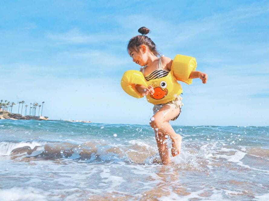 A little girl running in the ocean wearing a yellow life jacket.