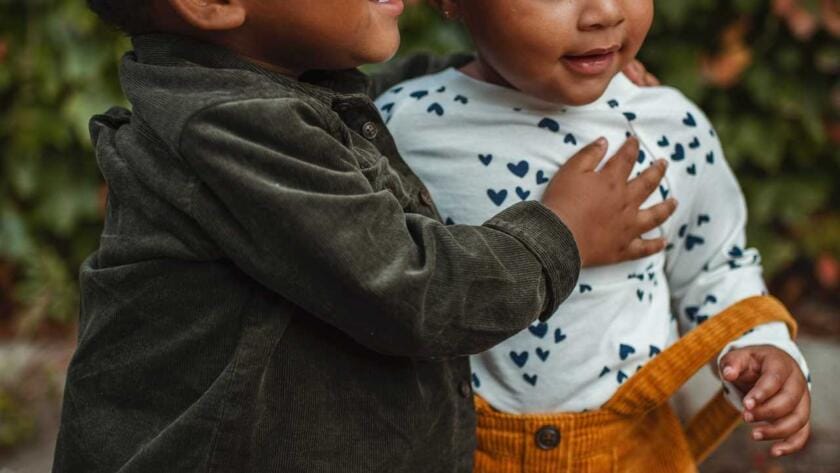 Two black children hugging each other in front of bushes.