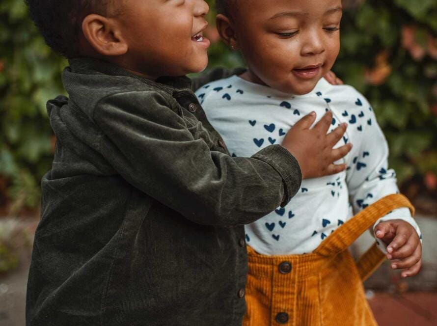 Two black children hugging each other in front of bushes.