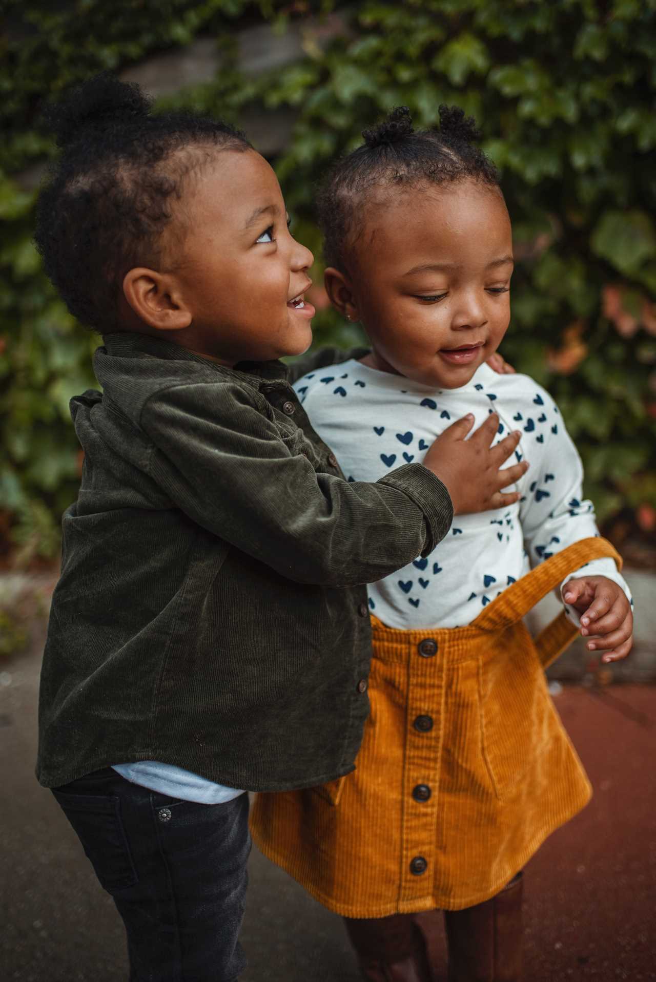 Two black children hugging each other in front of bushes.