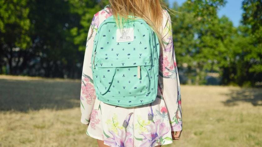 A girl in a floral dress is standing in a field with a green backpack.