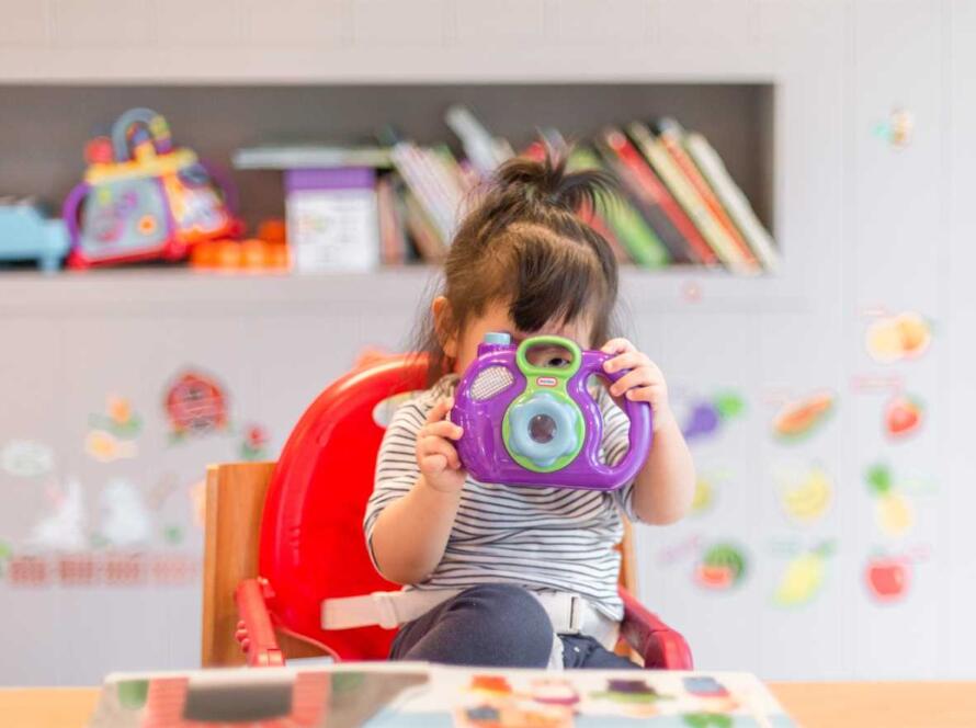 A little girl in a high chair taking a picture with a camera.