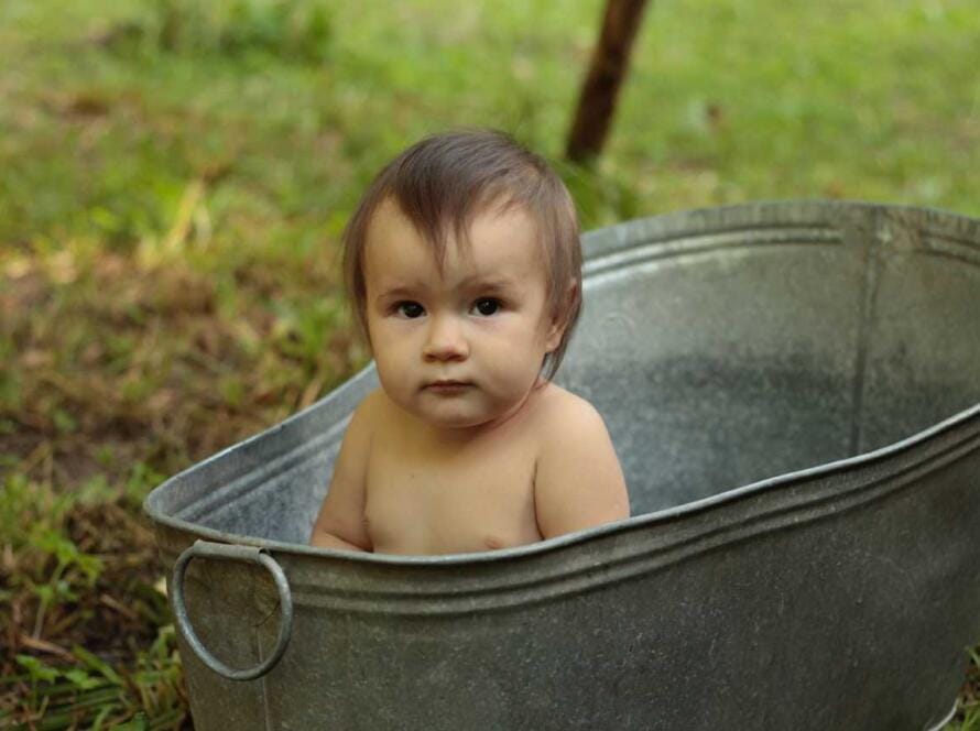 A baby is sitting in a metal tub in the grass.