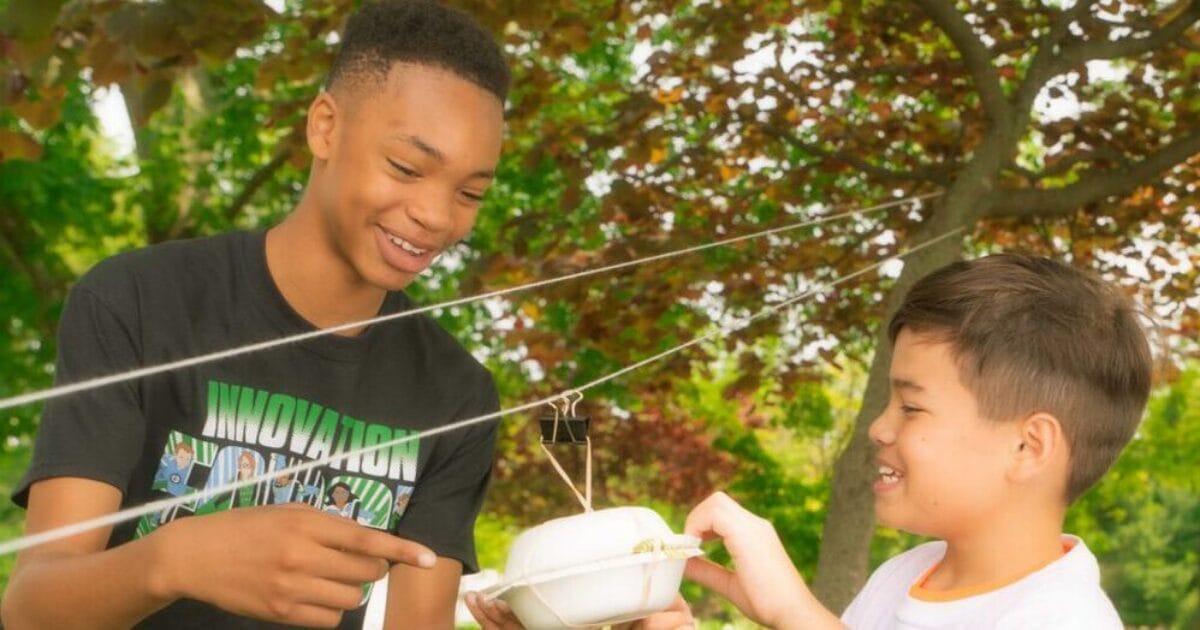 Two boys engaging in an outdoor science experiment with strings and a foam container.